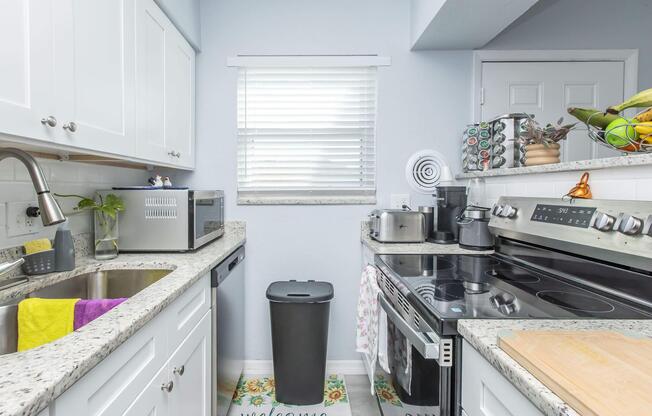 A modern kitchen featuring white cabinetry, a stainless steel stove, and a sink with a granite countertop. There is a trash can, kitchen utensils, and a decorative welcome mat on the floor. A window with blinds allows natural light to fill the space, enhancing the clean and tidy appearance.