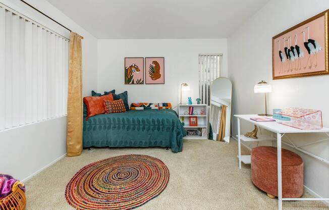A cozy bedroom featuring a turquoise bedspread with decorative pillows, a round woven rug, and two framed animal prints on the wall. A small desk with a lamp and a pink ottoman are present, along with a standing mirror and a shelf displaying decorative items. Natural light comes through window blinds.