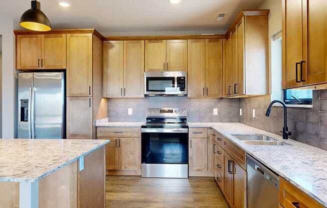 a kitchen with light brown wooden cabinets, stainless steel appliances, a tile backsplash, a large central island, and quartz countertops