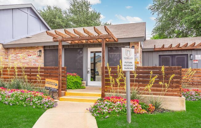 A welcoming entrance to an apartment complex featuring a wooden pergola, colorful flowers, and a sign for future resident parking. The building has a combination of brick and wooden elements, with large windows and a well-maintained lawn. Lush greenery and landscaping enhance the inviting atmosphere.