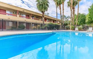 View of a sparkling blue swimming pool surrounded by a black fence, with palm trees in the background. An apartment building with a cream-colored exterior and red accents is visible in the background, reflecting in the water. The sky is partly cloudy, adding to the serene atmosphere.