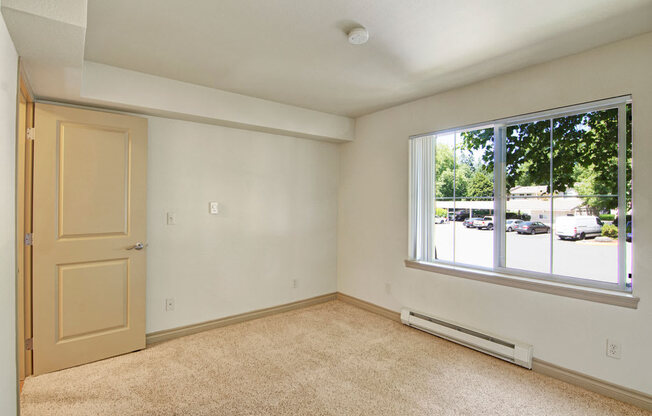 A neutral bedroom here at Asbury Park featuring plush carpet flooring, soft-toned walls, and a wide window with vertical blinds that brings in natural light and offers views of the community parking area, complemented by a simple door entry, clean trim details, and a calm layout ideal for rest and personalization.