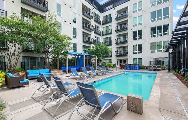 A landscaped outdoor pool courtyard here at Blue and Lime featuring a rectangular swimming pool surrounded by blue lounge chairs, shaded seating areas, and modern patio furniture. Multi-story apartment buildings with balconies frame the space, while trees and planters add greenery to the relaxing communal setting.