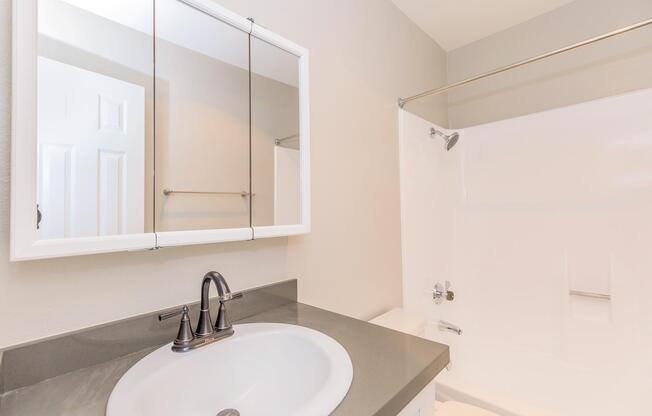 A clean, modern bathroom featuring a double-mirror above a circular sink with a sleek faucet. The shower area has a white tub and a clear shower curtain rod, while a light-colored wall provides a neutral backdrop. A door is slightly visible on the left side.