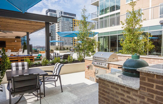 An outdoor grilling and dining area with a four-seat patio table, traditional stainless steel grill, and a Big Green Egg charcoal and ceramic grill. The prep spaces have granite countertops.