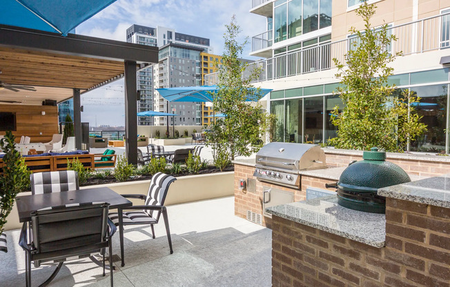 An outdoor grilling and dining area with a four-seat patio table, traditional stainless steel grill, and a Big Green Egg charcoal and ceramic grill. The prep spaces have granite countertops.