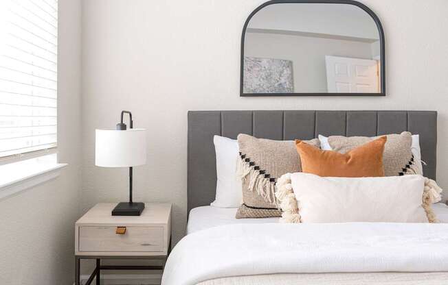A bed with a white comforter and a grey headboard with a white nightstand and a lamp on it.
