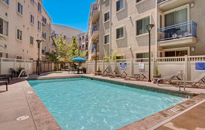 A pool courtyard here at Bridges with a rectangular pool, beige apartment buildings, blue balcony railings, tan deck, and lounge chairs, creating a bright, inviting outdoor amenity space.