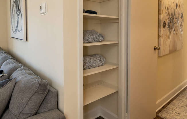 A grey couch sits next to a white wall with a shelf and a painting on it.