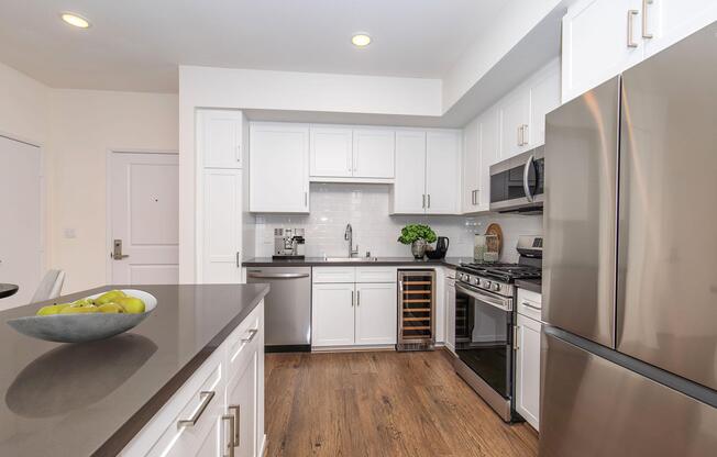 Modern kitchen featuring white cabinetry, stainless steel appliances, including a gas stove and refrigerator, a sleek countertop, and a wine rack. The space is well-lit with overhead lighting and includes a bowl of green apples on the counter, creating a clean and inviting atmosphere.