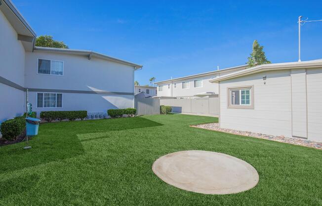 A landscaped outdoor area featuring a well-maintained green lawn, surrounded by two buildings. There is a circular concrete slab on the ground, with shrubs and a fence in the background, under a clear blue sky.