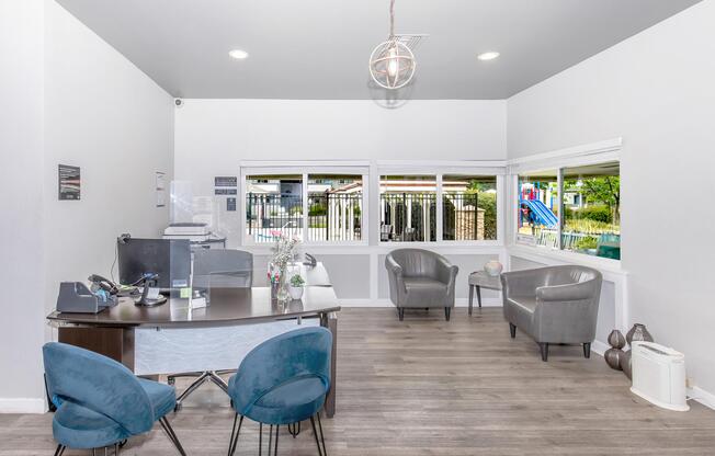 A bright and modern office space featuring a wooden desk, a computer, and comfortable seating arrangements including two gray chairs. Large windows allow natural light to flood the room, offering a view of an outdoor area with a pool and playground equipment visible in the background.