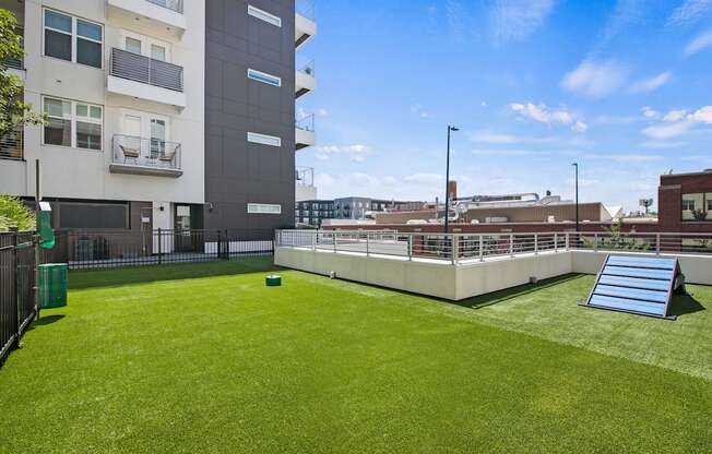 A large grassy area in front of a building with a metal fence and a small green trash can.
