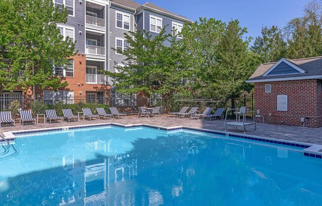 A clear blue swimming pool surrounded by lounge chairs and trees, with a background of a multi-story apartment building featuring balconies. The area is well-lit and inviting, perfect for relaxation and leisure.