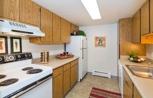 A functional kitchen here at Eagle Ridge showcasing warm wood cabinetry, laminate countertops, a full appliance setup with a refrigerator and range, overhead lighting, neutral vinyl flooring, a stainless sink with ample counter space, and simple wall décor that adds a cozy, practical feel to the space.