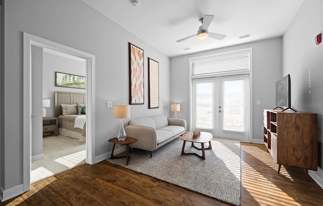 A spacious living room with hardwood-style flooring, double doors to a patio, and a ceiling fan near an doorway to a bedroom.