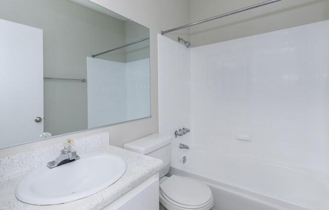 A clean, modern bathroom featuring a white bathtub, a toilet, a sink with a simple countertop, and a large mirror. The walls are painted in a neutral tone, and there is a shower curtain on the side. The overall ambiance is bright and uncluttered.