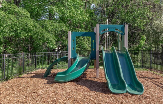 Two green slides on a playground structure surrounded by a wooden mulch surface and a fenced area, with lush green trees in the background. The playground set is designed for children, featuring a modern design and a safe play environment.