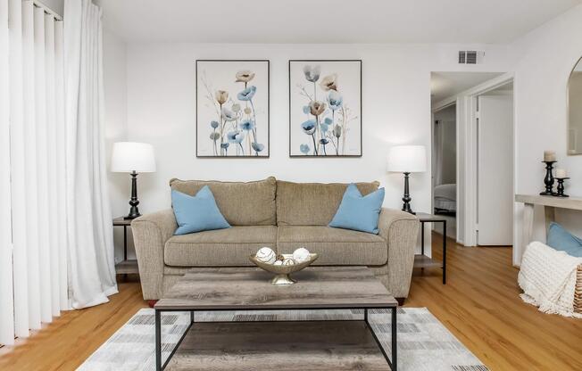 A cozy living room featuring a beige sofa with blue accent pillows, a wooden coffee table with decorative items, and light-colored walls. There are two lamps on either side of the sofa and artwork of flowers in soft colors hanging above. Natural light brightens the space, creating a welcoming atmosphere.
