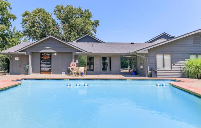 A bright and inviting outdoor swimming pool area surrounded by a wooden deck. In the background, there is a modern building with large windows and a sign. Lounge chairs are placed near the pool, and there are trees and greenery in the surrounding landscape. Clear blue skies overhead complete the scene.