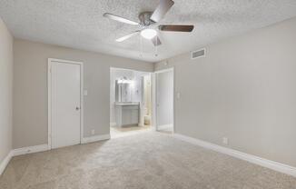 A spacious, lightly colored bedroom featuring beige carpet, a ceiling fan with wooden blades, and a doorway leading to a bathroom area in the background. The walls are painted in a soft tone, and there is a closed white door on the left. Natural light highlights the room's neutral decor.