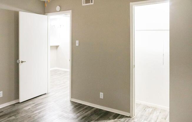 A brightly lit room with light gray walls and new laminate flooring. Two doors are visible: one leads to a closet space, while the other opens to another room. The area is clean and minimalistic, creating a spacious feel.