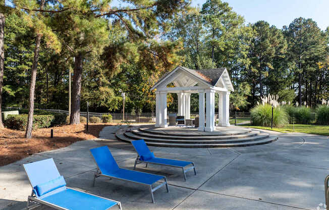 A gazebo is surrounded by trees and has two blue lounge chairs in front of it.