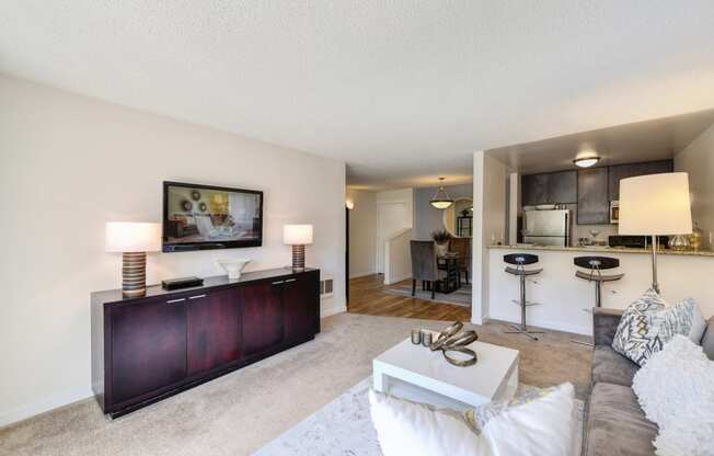 Model Living room with gray couch and white chair.  Kitchen and counter with bar stools is visible from the living room.