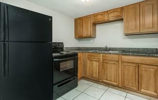A small kitchen featuring dark wood cabinets, a black refrigerator, and a black stove with an oven. The countertop is made of granite, and the floor is tiled. There is a single sink in the corner, with a light fixture above providing illumination. The overall space appears clean and well-maintained.