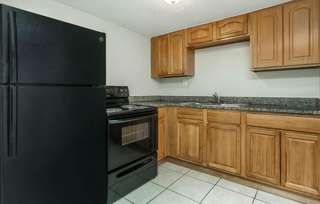 A small kitchen featuring dark wood cabinets, a black refrigerator, and a black stove with an oven. The countertop is made of granite, and the floor is tiled. There is a single sink in the corner, with a light fixture above providing illumination. The overall space appears clean and well-maintained.