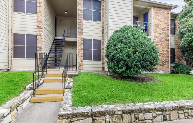 Exterior view of an apartment building featuring a set of stairs leading up to the entrance, surrounded by well-maintained grassy areas and a rounded bush.
