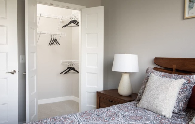 A bedroom with a bed, a nightstand with a lamp, and a closet with clothes hanging.