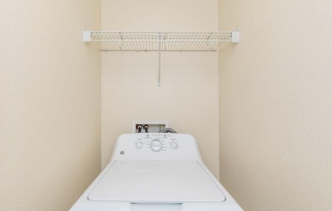 A white washing machine in a small laundry space with light beige walls. Above the machine, there is a metal wire shelf for storage. The area is clean and organized, suggesting a functional home laundry setup.