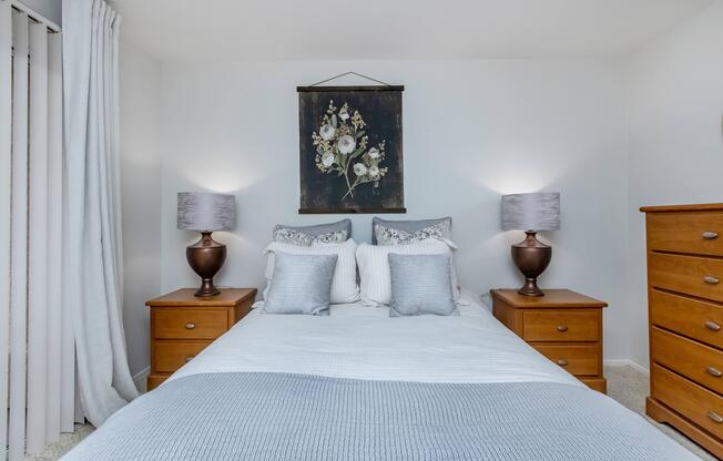 A cozy bedroom featuring a double bed with decorative pillows, two wooden nightstands with lamps, and a floral wall art above the bed. Soft gray bedding complements the decor, and light curtains drape over a sliding door, creating a serene atmosphere. Natural light enhances the inviting space.