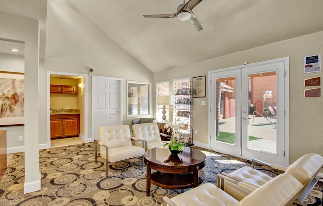 A welcoming resident lounge here at Aspire Pinnacle Park featuring comfortable cushioned seating arranged around a round wood coffee table, patterned carpet flooring, and large glass doors that open to the pool area. A refreshment nook with cabinetry and a sink, brochure display rack, and soft lighting create a bright, inviting space to relax or meet with neighbors.