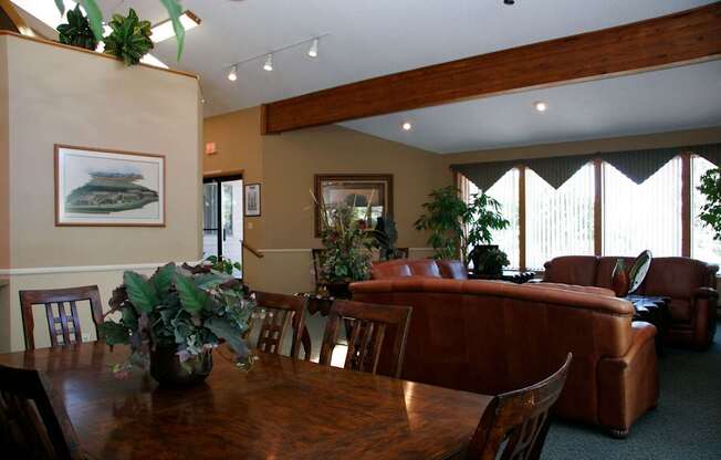 A welcoming community gathering space here at Eagle Ridge featuring a wooden dining table with matching chairs in the foreground, brown leather sofas arranged for conversation, exposed wood ceiling beams, neutral walls with framed artwork, tall windows with vertical blinds letting in natural light, and greenery accents that create a warm, comfortable atmosphere.