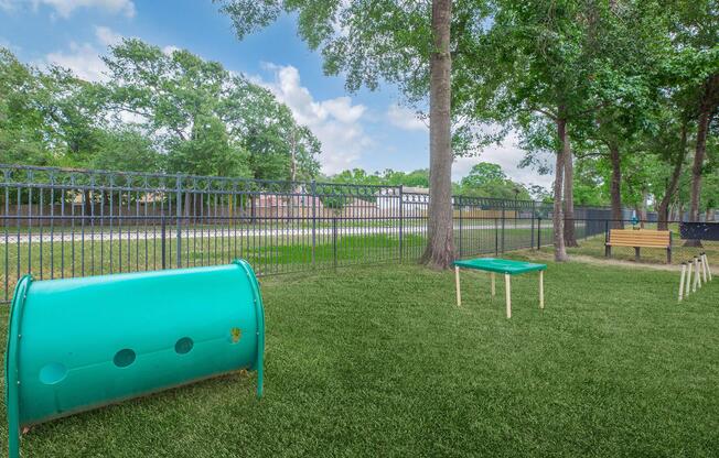 A green play area featuring a tunnel and a table on artificial grass, surrounded by trees and a black fence. In the background, thereâs a view of a road and houses. The setting appears inviting and suitable for outdoor activities.