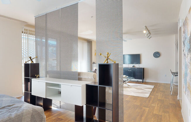 A modern open bedroom here at Rio Paradiso featuring a stylish shelving divider with textured panel screens that separate the sleeping area from the living space, complemented by wood-style flooring, soft natural light from large windows with vertical blinds, contemporary décor accents, and a clear view into the living room with seating, media console, and wall art.