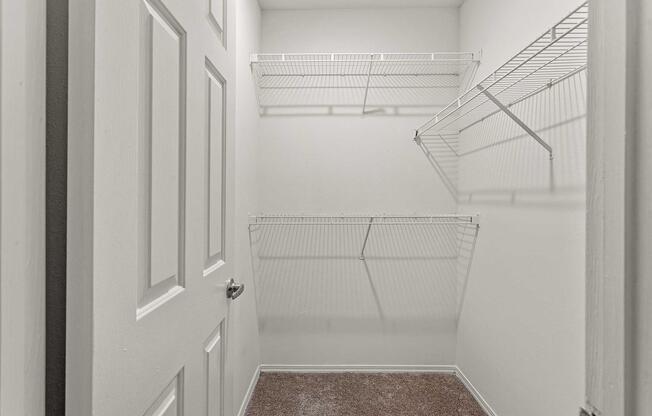 Empty closet with white walls and wire shelving. The closet features two wire racks for storage and a carpeted floor. The door is closed, and the lighting is neutral, highlighting the simplicity of the space.