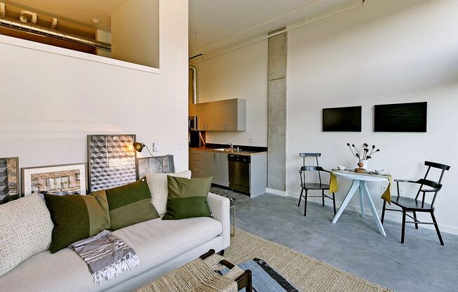 Living room with partial wall behind couch, leading to loft area. Staged with a couch, coffee table, rug and chair. Small dining area at right, with kitchen behind.at 19th & Mercer, Seattle Washington