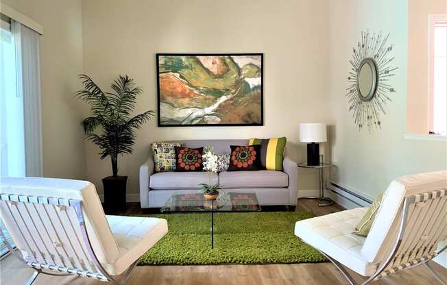2 bedroom model living room with a tan couch, 2 white accent chairs, glass coffee table, green accent rug, plants, lamp and wall decorations