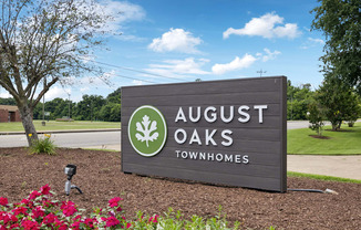 A sign for August Oaks Townhomes is displayed in front of a tree and flowers.