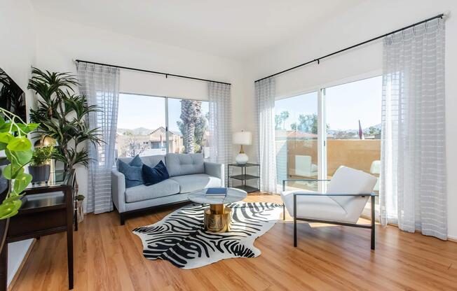 A bright and airy living room featuring a light-colored sofa, a glass coffee table, and tall windows with sheer curtains. Potted plants add a touch of greenery, while light wood flooring complements the modern decor. The view outside includes a sunny landscape, enhancing the inviting atmosphere.