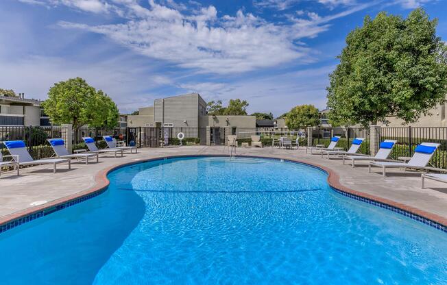 A clear, inviting swimming pool surrounded by lounge chairs with blue cushions. Lush green trees are nearby, and modern apartment buildings can be seen in the background. The sky is bright blue with scattered clouds, creating a relaxing and sunny atmosphere perfect for leisure.