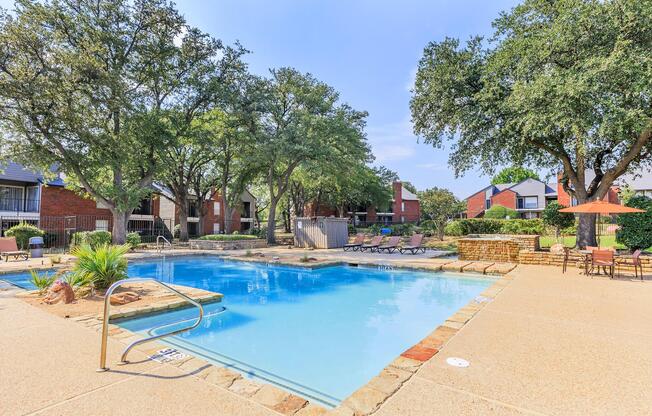 A serene outdoor swimming pool surrounded by lush green trees and lounge chairs. The pool area features a stone edge, with patio tables and umbrellas for shade, creating a relaxing atmosphere in a residential setting. Bright blue skies enhance the inviting scene.