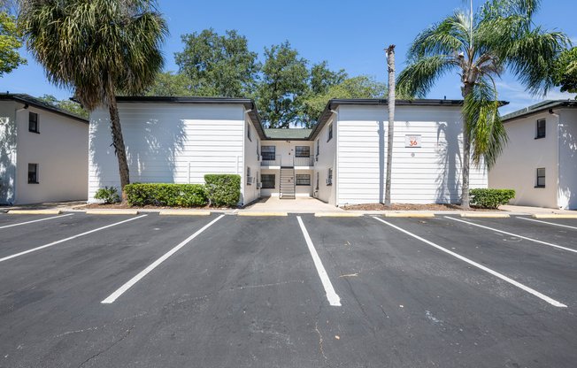 Inviting apartment exterior with modern white facade, surrounded by lush greenery and palm trees, featuring ample parking space.