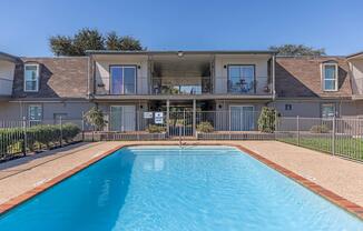 A clear blue swimming pool in the foreground, surrounded by a tidy stone deck. In the background, a two-story apartment building with balconies overlooks the pool area. Lush greenery and well-maintained plants decorate the space, creating a welcoming atmosphere under a bright blue sky.