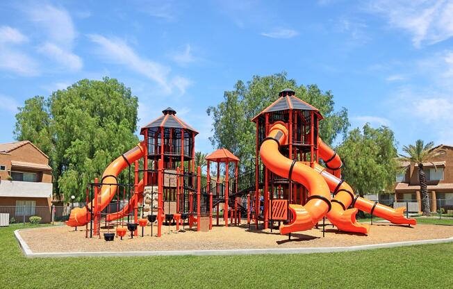 A colorful playground featuring two large orange slides, climbing structures, and play areas. The playground is set in a grassy area with trees and residential buildings in the background under a bright blue sky.