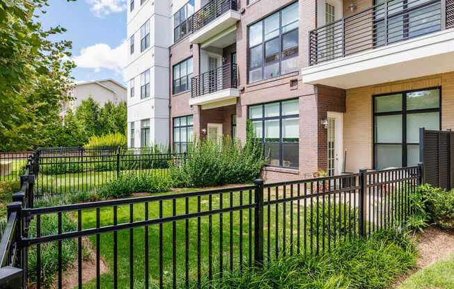 A black metal fence surrounds a grassy area in front of a multi-story apartment building.
