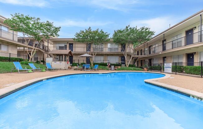 A clear blue swimming pool surrounded by lounge chairs and shade structures, located in a courtyard area flanked by two-story apartment buildings with balconies. Lush green trees are visible in the background under a bright blue sky.